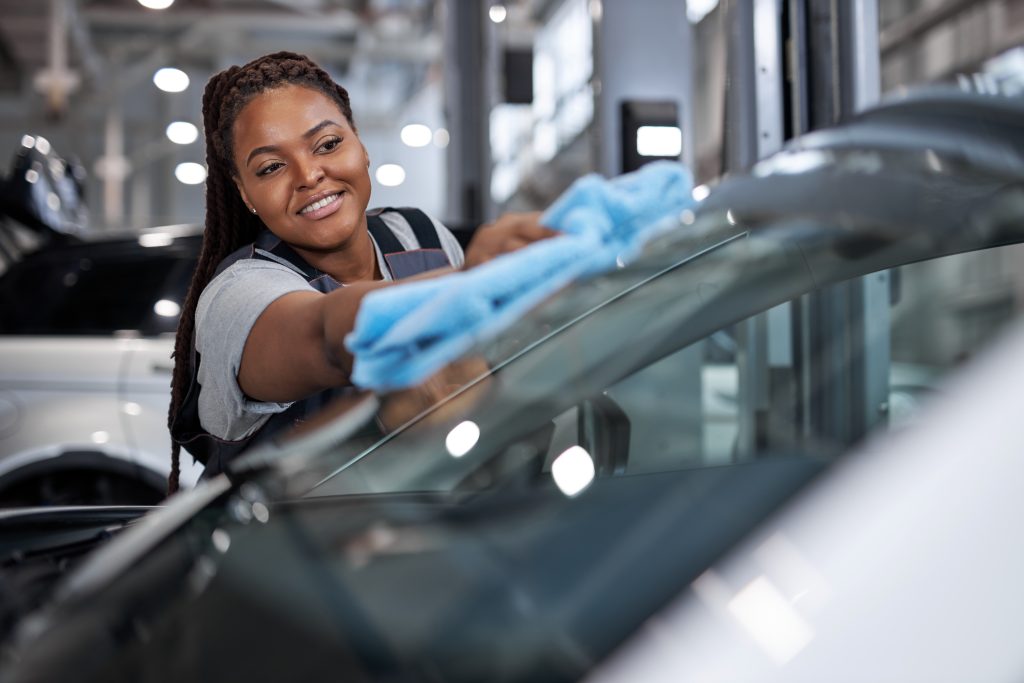 A female mechanic with braids smiling while wiping a car windshield.