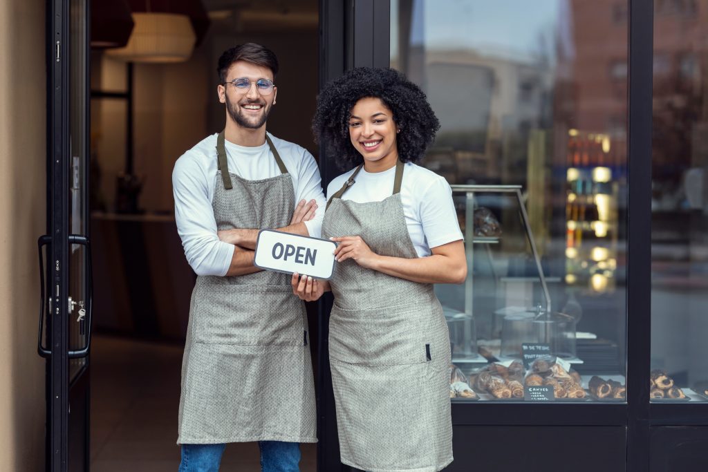 Two smiling bakery owners holding an "OPEN" sign at their shop entrance.